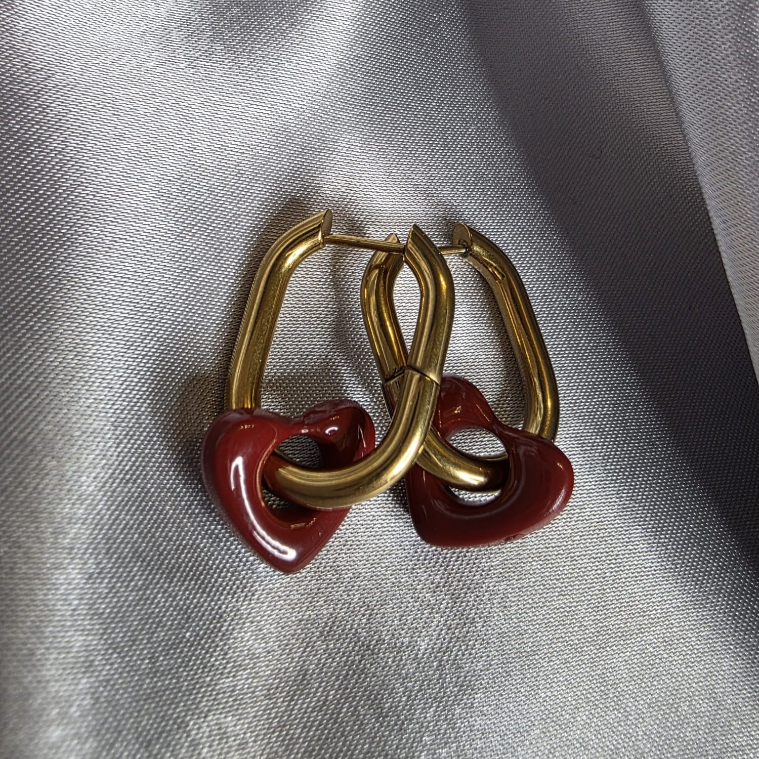 Gold hoop earrings with red heart-shaped charms on a gray fabric background